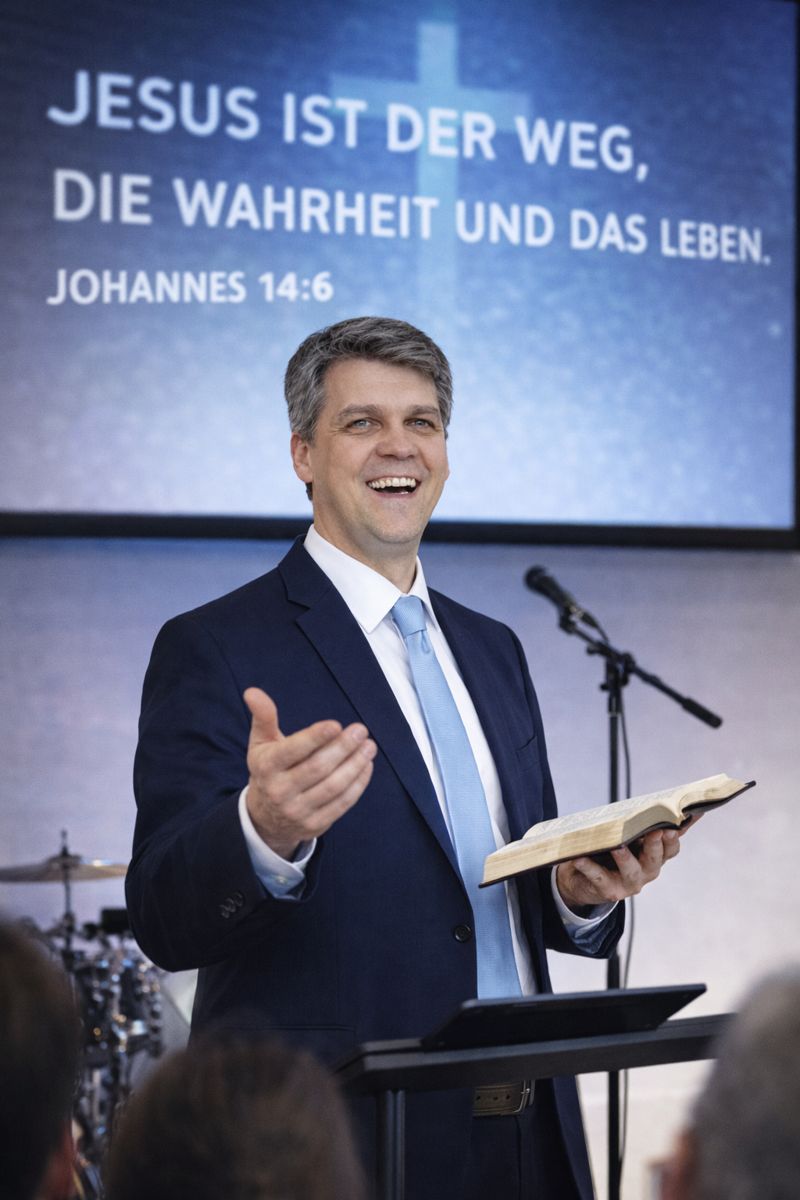 Pastor Christian steht im Anzug und hält eine geöffnete Bibel, umgeben von Menschen in einer Gemeinde